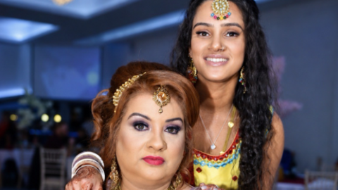 A woman with long dark hair, jewellery, and a yellow dress with her arm on the shoulder of a woman with ginger hair and jewellery