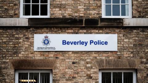 A sign which reads 'Beverley Police' with a white background positioned on a sandy brick walled building with four windows. A police logo can be seen to the left of the sign.