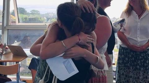 Two young women hug after getting exam results. One is holding an envelope.