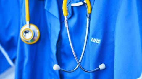 A close up of a healthcare working in blue scrubs with the white and blue NHS logo on the breast. There is a silver and yellow stethoscope around the person's neck