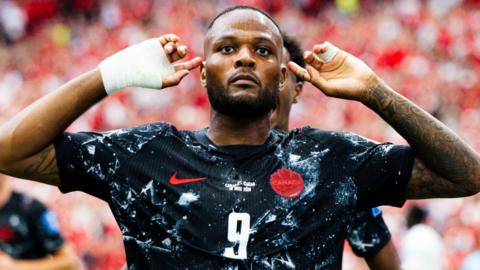 Cyle Larin, wearing Canada's black away strip with white patterns on it, celebrates scoring at the World Cup against Qatar by putting his hands either side of his head and pointing to his ears with his index fingers