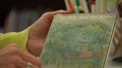 A close up of the book The Boy and the River by Henri Bosco. It is a hardback edition with a worn dust jacket, showing a watercolour painting of a two little boys in a boat.