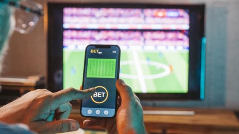Man watching football match on TV while making a bet on a gambling app on phone