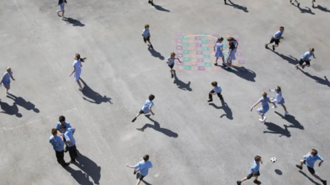 More than a dozen boys and girls play in a playground in sunshine. The aerial photo show children in a blue uniform playing or chatting together in small groups or running around.