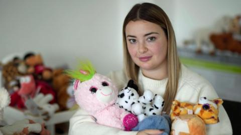 Hollie Edridge is looking at the camera with a calm expression. She has shoulder length blonde hair and is holding an armful of cuddly toys. There are more in the background.