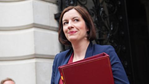 Woman with short brown bob in navy blazer carrying red folder walking out of white stoned government building 
