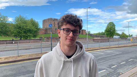 Alfie Goodwin standing on a pavement with Carlisle Castle in the background. He has short brown curly hair and blue eyes. He is wearing glasses with a dark rim and a cream hoodie.