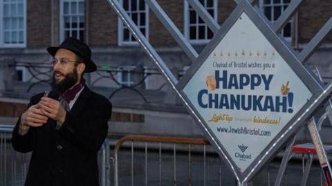 Rabbi Mendy is pictured standing in front of a display that says 'Happy Chanukkah' in College Green, Bristol City Centre. He is wearing a black wide-brimmed hat and a long, black coat. He has a bushy black beard and is wearing glasses. There is a building and metal fencing behind him.