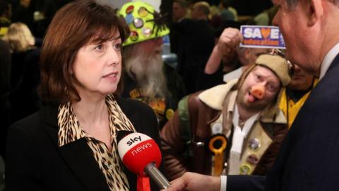 Deputy Labour Party leader Lucy Powell talking into a red microphone - as The Official Monster Raving Loony Party candidate reacts in the background
