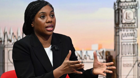 Kemi Badenoch sitting in a red chair is looking at the camera and wearing a white blouse and black suit jacket.