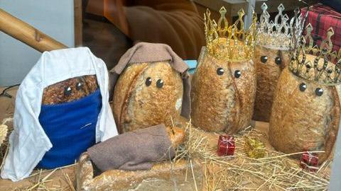 A Nativity scene made out of bread arranged in a bakery window. The scene features three sourdough loaves wearing crowns for the three kings, sourdough loaves Mary and Joseph wearing cloth, and a baguette wrapped in cloth sleeping on a rye loaf for baby Jesus.