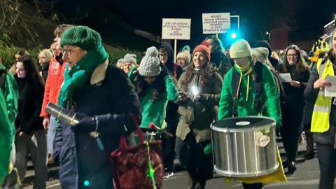 A group of people, mostly women wearing green, walk down a road during  a march at night, some with signs and drums.