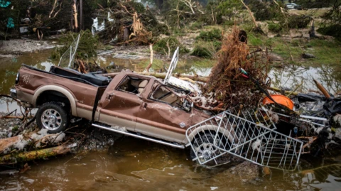 Brown pick up truck with smashed windows and partially crushed-in doors is buried under debris and water after flooding in Texas