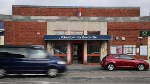 Pokesdown Station is a small red-brick building with a blue name sign above some glass doors. It is on a main road, and cars are driving past it.