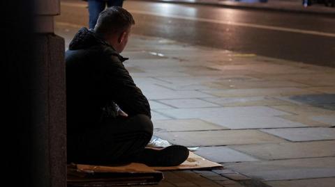 A homeless man sits on a piece of cardboard on a street at night, he is wearing dark clothing and facing away from the camera. He has short dark hair.