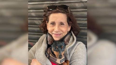 Ruth Myers smiling at the camera, with sunglasses resting on her head. She has short brown hair is holding a small black dog and wearing a grey hoodie.