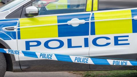 A police car with yellow and blue hi-vis decals parked on a grassy bank behind a strip of blue and white police cordon tape.