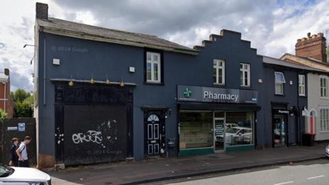 A stretch of grey buildings are on one side of the road with one having a pharmacy sign above it with another boarded up with some graffiti on it.
