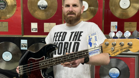 Auctioneer Luke Hobbs from Gardiner Houlgate stands facing the camera holding a black bass guitar which was played by Chris Cross at Live Aid. Mr Hobbs has a beard and is wearing a white T-shirt with "Feed the World" in black letters on it