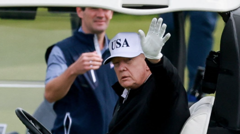 Donald Trump is wearing a white golf cap and glove, and a black top. He is waving as he sits behind the wheel of a golf bubby