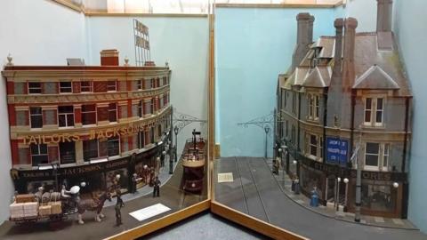 Two intricate and realistic models of buildings. The one on the left has a curved redbrick facade and says "Jacksons Corner" on it, with a tram and a horse and cart ouside. The one on the right says "Duke Street stores" on it and is made of grey brick.
