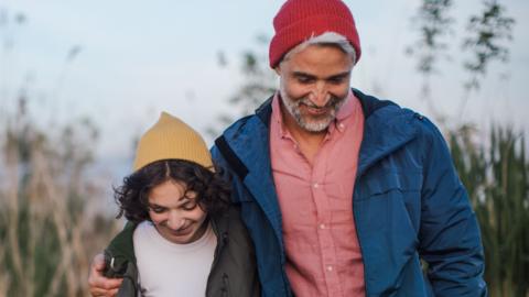 A father and daughter walk through a tall grasses, both are looking down and smiling, and are wearing colourful hats.