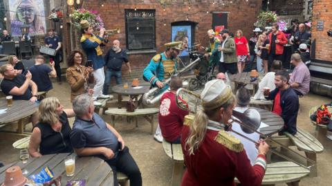 Band performing to pub goers in the Tamworth Tap Stop pub garden. The band wear blue, dark blue or burgundy jackets with gold trim on their shoulders and play brass instruments. One plays while standing on a wooden bench. The band is watched by pub goers, some who are taking footage on their phones. 