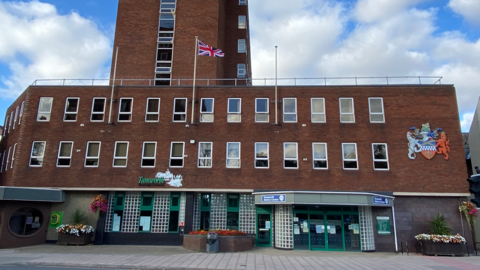 A building with a green sign saying Tamworth above green windows, a county crest mounted on the brickwork and a Union Jack flag flying from a pole. The building has three stories, with a taller tower section.