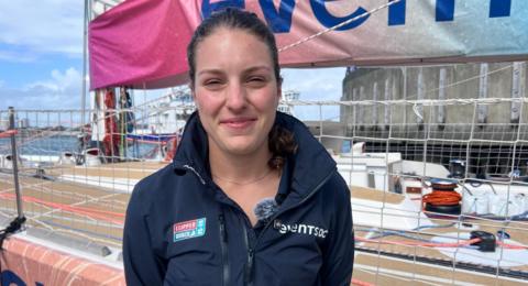 Ella Hebron smiling for the camera. She is standing on the quayside with one of the yachts behind her. She has tied back brown hair and is wearing a navy blue sailing jacket.