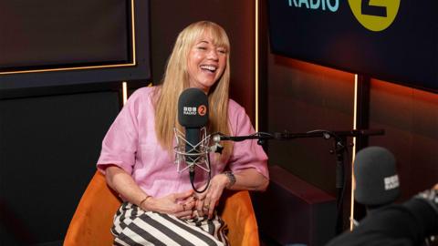 Sara Cox dressed in a pink shirt and stripey trousers, sitting in an orange chair speaking into a Radio 2 microphone, smiling, laughing