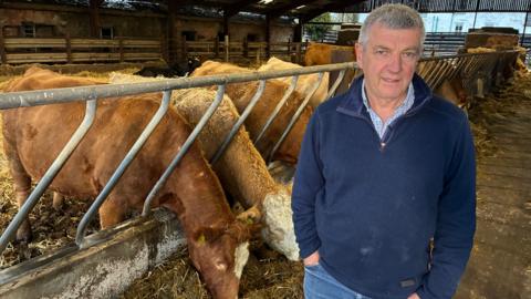 A man stands in a large shed in front of penned cattle, which are grazing on hay. The man has short grey hair and is wearing blue jeans and a dark blue fleece - he stands with his hands in his pockets looking into the camera.