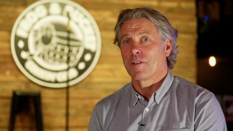 John Bishop is talking in this screenshot from the interview, with the blurred out Frog and Bucket logo and the stage and microphone seen behind him.