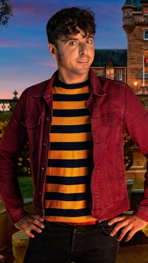 James Baker poses for his Traitors promotional image. He wears a red and yellow striped top with a red denim jacket over the top. He has short dark brown hair which is curly on top.