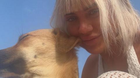 A photograph of the head and shoulders of a woman with short blonde hair on the right, with her arm around a light-haired dog.