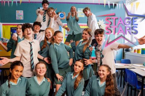 Stage Stars students sitting together in a classroom, smiling - the Stage Stars logo in the top right