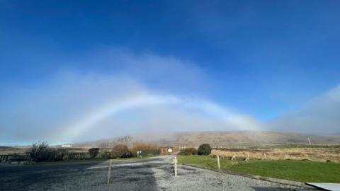 A fog bow sits in a bright blue sky arching over fields with a gravel road in the foreground