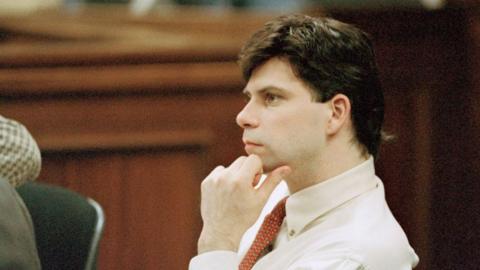 Archive image of Lyle Menendez in court in the 1990s as a young man, listening with his hand on his chin, wearing a white shirt and patterned red tie. 