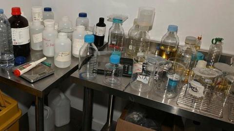 A laboratory workspace with stainless steel benches covered in assorted glassware and chemical containers. Numerous clear and opaque plastic bottles, reagent bottles, beakers, measuring vessels and sample containers are arranged across the work surfaces, some containing amber-coloured liquids. A digital calculator and laboratory tools are visible among the equipment, while larger storage containers are stored underneath the benches. The image depicts a busy laboratory preparation or testing area with a variety of chemicals and scientific apparatus.