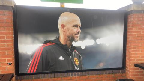 A poster of Erik Ten Hag outside Old Trafford by a red brick seating area 