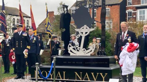 Royal Navy personnel gather round the statue holding flags.  The statue sits on a plinth that says Royal Navy in large gold letters.  There are three figures in 2D black metal surrounding a large silver anchor.