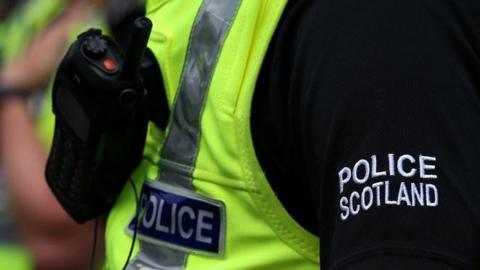 Police Scotland uniform, yellow bib with work police, and Police Scotland in bold white letters on a black sleeve.
