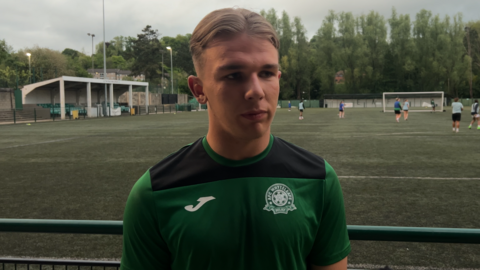 A blonde boy with a centre parting. He is wearing a green AFC Whyteleafe training shirt and is standing in front of a 3G pitch.