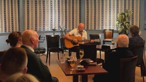 A man plays guitar at the back of a room. A crowd of people are sat at tables with drinks watching. The room has a brown wooden floor and patterned blue and white curtains.