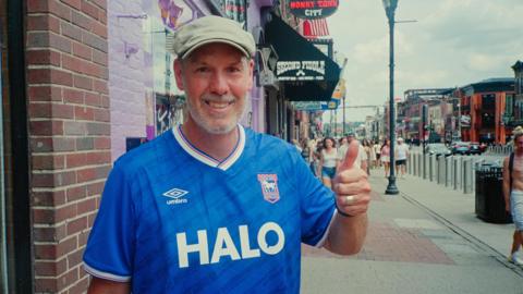 A man standing outside on a pavement beside a road. He is wearing a cream coloured flat cap and a blue football shirt. He is smiling at the camera and has one of his arms raised up, with his thumb up.