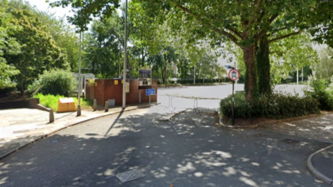 The approach to a car park, with barriers across the entrance, and trees overhanging the area