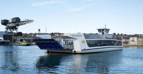 White chain ferry mid crossing on a stretch of river.  To the right on the shore are rows of buildings to the left of the ferry is a large crane.
