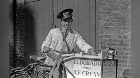 A black and white image dating from the inter-war period of an ice cream seller with a bicycle. He is wearing a white overall and a peaked cap and standing, leaning on his pedal bike, which has the ice cream container on the front labelled "Eldorado Pure Ice Cream.
