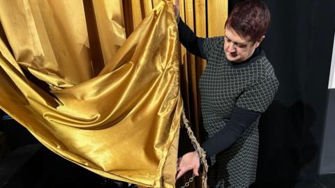 A woman with short, red hair and a black and white patterned dress holding up a huge gold screen curtain which is damaged and stained at the bottom.