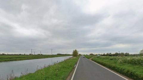 A road, with water to the left and two lanes of traffic. Cars are on it with grass either side of the road and pylons and buildings to the right, in the distance. 