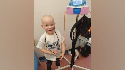 Jaxson in a hospital room with no hair, a tube coming out of his nose, and attached to a small machine on a stand. He is wearing a t-shirt saying 'Fighter'